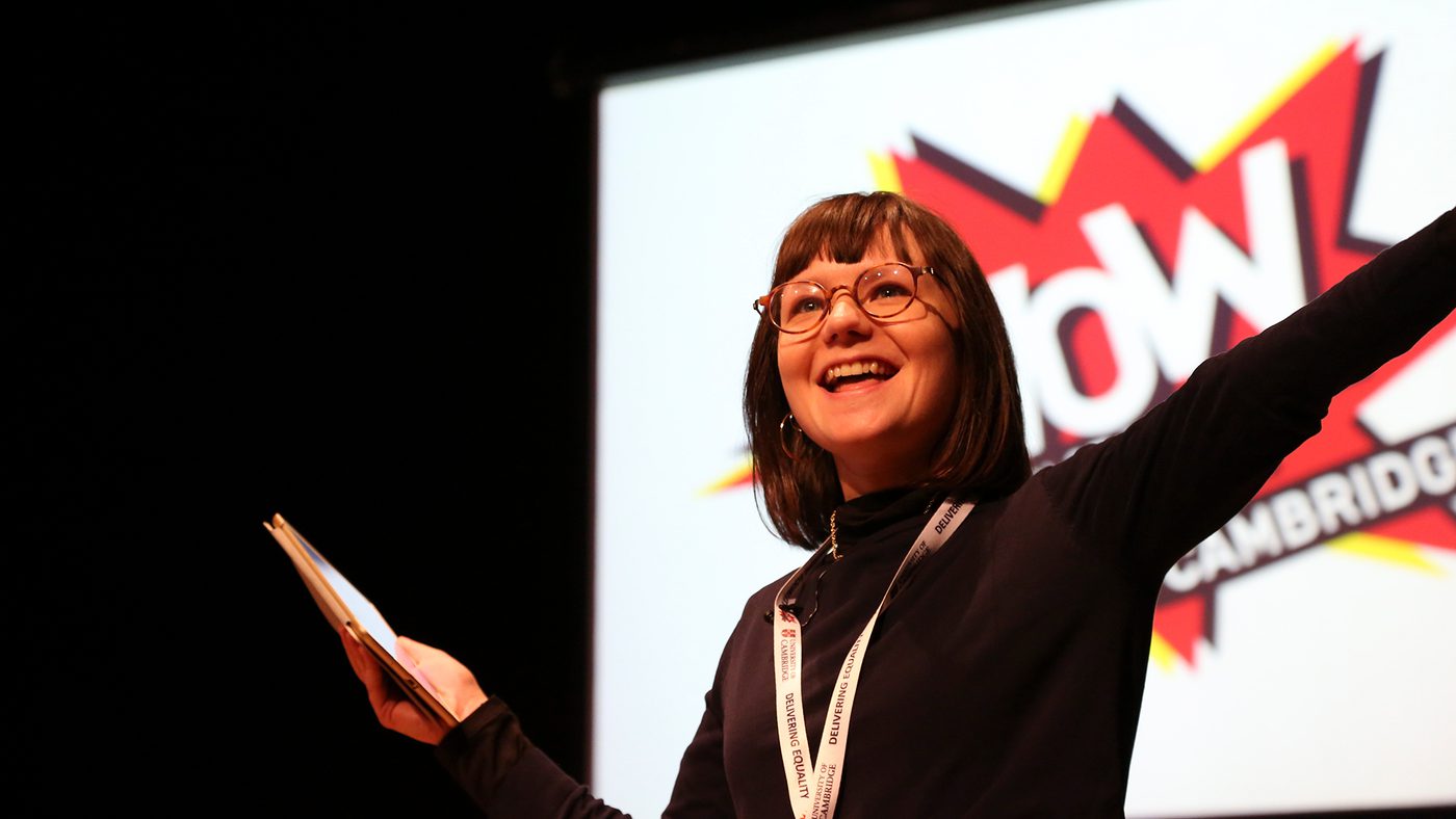 A woman with glasses smiles enthusiastically on stage, holding a notepad. Behind her is a "WOW Cambridge" sign.