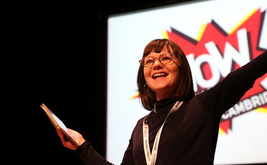 A woman with glasses smiles enthusiastically on stage, holding a notepad. Behind her is a "WOW Cambridge" sign.