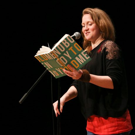 A woman stands on stage, reading from a book into a microphone. A "WOW" event banner is visible beside her.