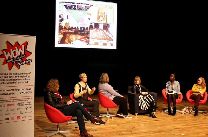 A panel of six people sits on a stage in red chairs under a projected presentation. A poster with "WOW" is on the left.