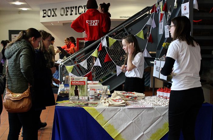 A busy box office area features a promotional table with brochures and gifts. People browse attentively, while staff in white shirts assist them.