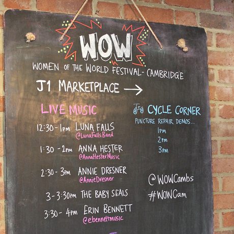 A black chalkboard sign on a brick wall lists event schedules for a festival. It includes live music times, artist names, and workshop details in colourful chalk.