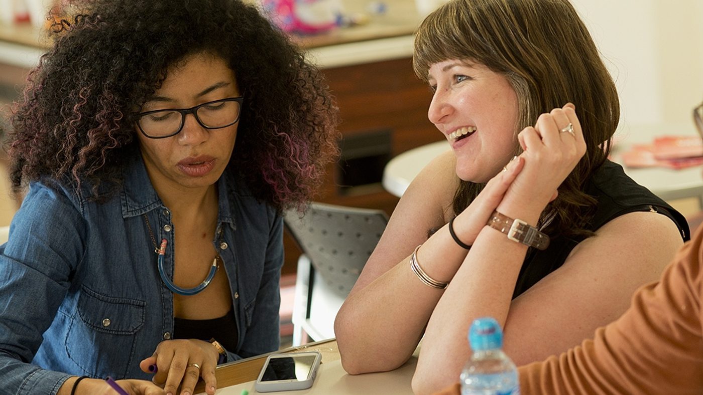 Two women sat at a table. One is concentrating hard and writing on a piece of paper, whilst the other is smiling and looking at something out of shot.