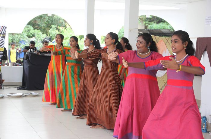 A group of young women performs a traditional dance, wearing colourful dresses and jewellry. They are in sync, with joyful expressions, and a backdrop of greenery.