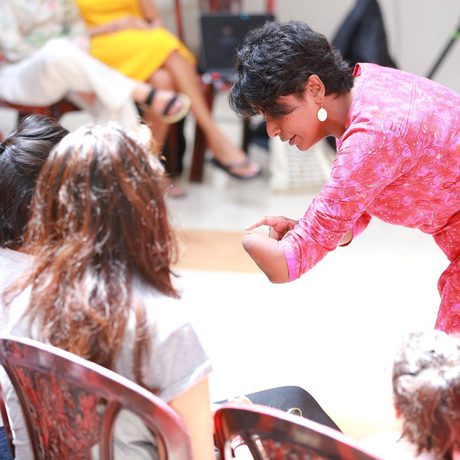 A woman in a pink top leans forward, engaging with seated individuals in a bright, informal setting.