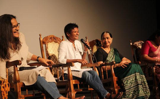 A panel of four women seated in ornate chairs on a stage, engaged in discussion. The atmosphere is lively, with smiles and casual attire.