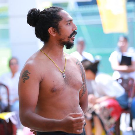 A shirtless man with a beard and bun stands outdoors. He wears a necklace and has a tattoo on his arm. Onlookers are blurred in the background.