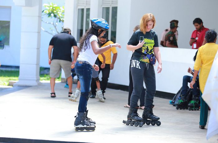 Two people rollerblading outdoors, concentrating and balancing. Others in the background observe.