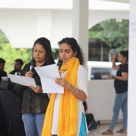 Two people stand indoors, holding microphones and reading from papers. They appear focused. Background has people and event posters.