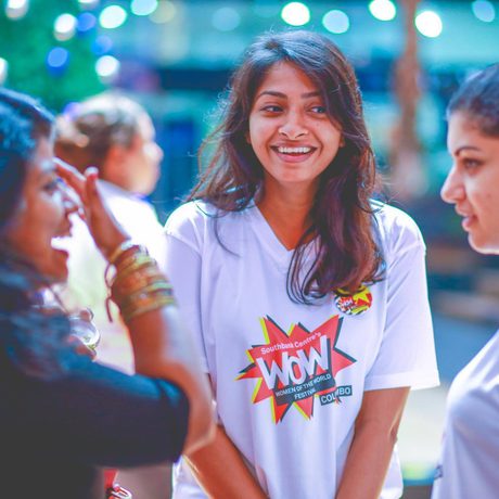 A group of four women in animated conversation at an outdoor gathering. One in a "WOW" t-shirt smiles brightly, creating a joyful and lively atmosphere.