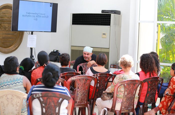 A small group of people sits in plastic chairs facing a presenter in a casual talk. A screen displays a presentation in a bright, airy room.