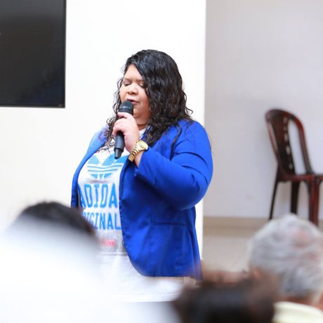 A woman in a blue blazer speaks into a microphone indoors, facing an audience. A poster is partially visible on the wall behind her.