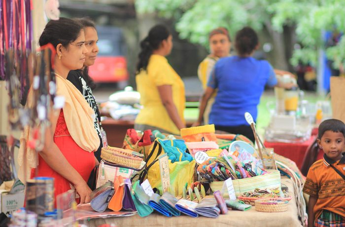 A vibrant market scene shows women behind a stall with colourful handcrafted items. In the background, blurred greenery and people.