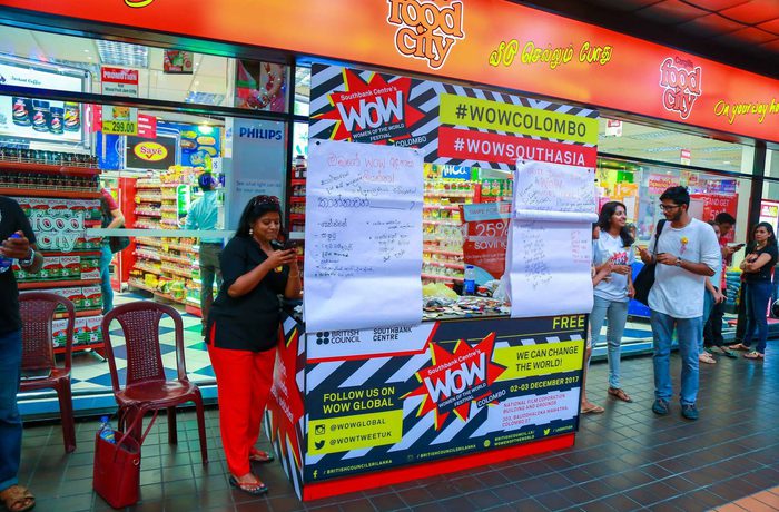 A lively storefront scene with people gathered around a colourful promotional booth displaying large posters.