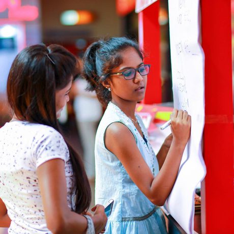 Two young women are engaged in discussion at an event. One writes on a board, while the other looks on.