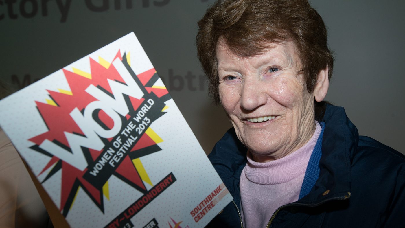 Woman smiling and holding a "Women of the World Festival 2013" pamphlet. She wears a blue jacket and is in front of a blurred grey background.