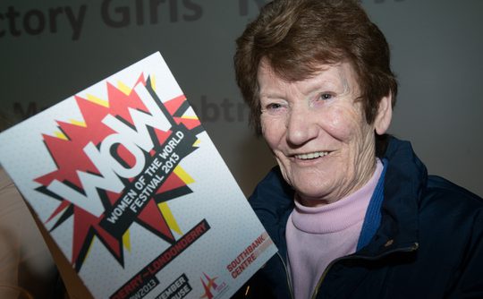 Woman smiling and holding a "Women of the World Festival 2013" pamphlet. She wears a blue jacket and is in front of a blurred grey background.