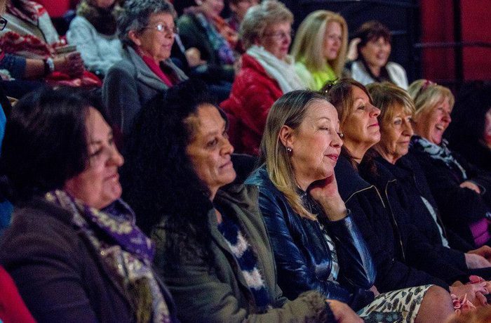An audience of diverse women, seated closely in a theatre, watch something attentively with a sense of interest and focus.
