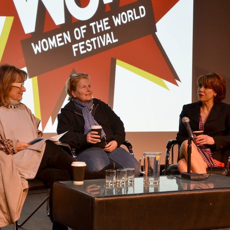 Three women sit on a stage panel at the "Women of the World Festival." There's a mic on the table. The setting is professional and conversational.