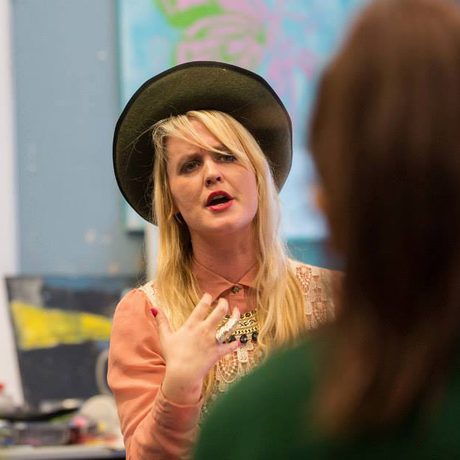 A woman wearing a hat and pink top speaks expressively to two seated people. Bright, colourful artwork is blurred in the background
