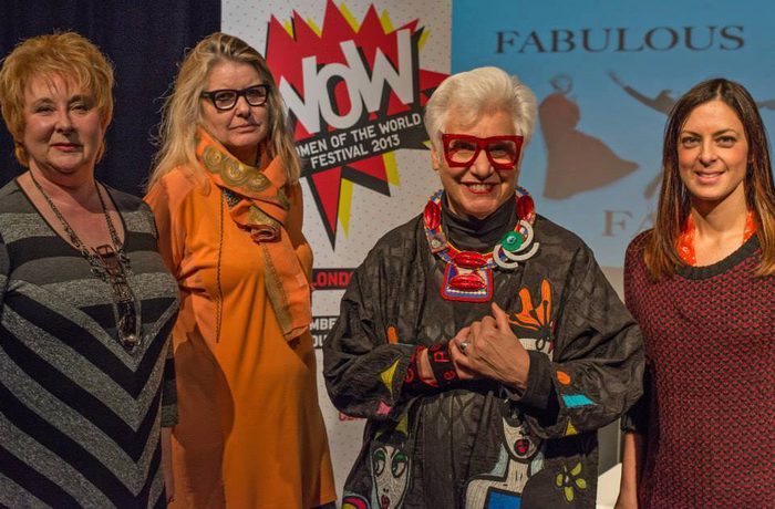 Four women stand together, smiling, at an event with a "Women of the World Festival 2015" sign. They appear confident and stylish.