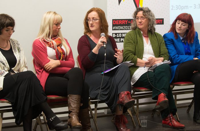 Five women sit on chairs in a panel discussion. They are attentively engaged, with one woman speaking into a microphone.