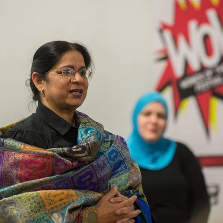 A woman in a colourful shawl speaks confidently at an event. A "WOW" banner is behind her. Another woman in a blue hijab listens intently.