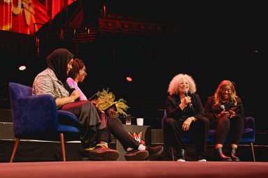 Four women sit on stage engaged in a lively discussion. Two hold microphones, and all seem relaxed and happy. The background features vibrant red decor.
