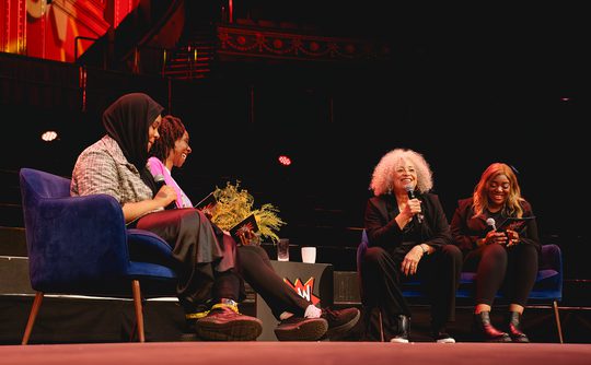Four women sit on stage engaged in a lively discussion. Two hold microphones, and all seem relaxed and happy. The background features vibrant red decor.