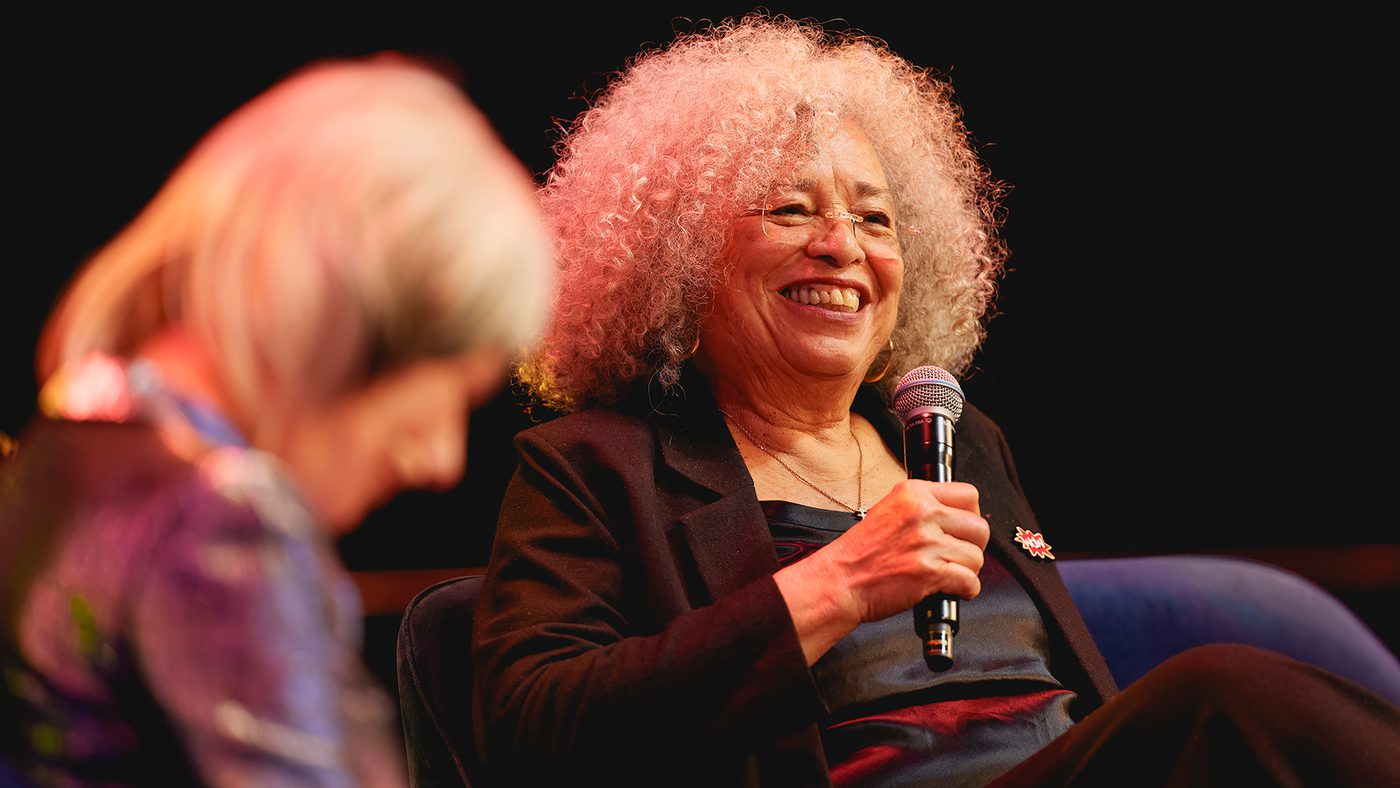 Angela Davis smiling and holding a microphone on stage. Another person in the foreground is slightly blurred. Warm, engaging atmosphere.