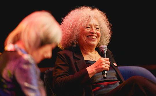 Angela Davis smiling and holding a microphone on stage. Another person in the foreground is slightly blurred. Warm, engaging atmosphere.