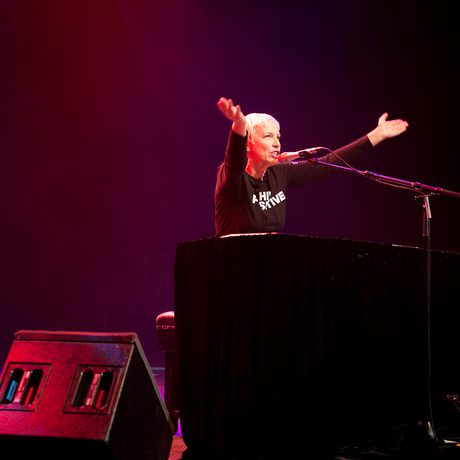Annie Lennox performs on stage at a piano under red lighting. Their arms are raised