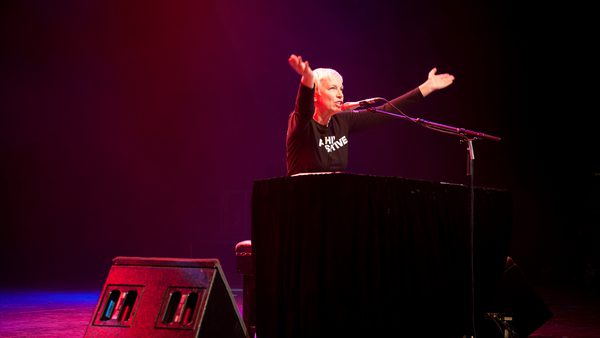 Annie Lennox performs on stage at a piano under red lighting. Their arms are raised