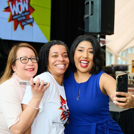 Three women smiling and taking a selfie at an event. The woman in blue holds the phone. A "Wow" poster is in the background