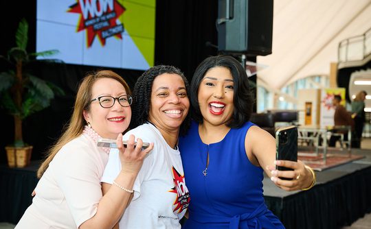 Three women smiling and taking a selfie at an event. The woman in blue holds the phone. A "Wow" poster is in the background