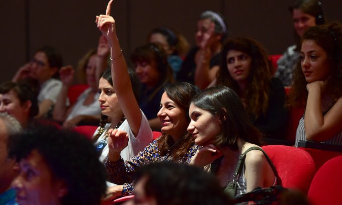 A diverse audience sits in a theatre with red seats, engaged in a presentation. A smiling woman raises her hand