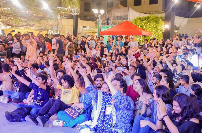 A vibrant, diverse crowd at an outdoor festival, sitting and cheering energetically.