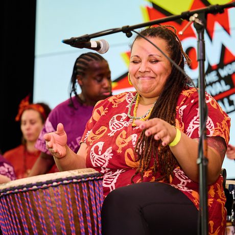 A group of diverse women joyfully play drums on stage. The central woman smiles with eyes closed. A banner in the background reads "WOW."