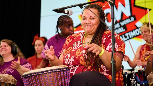A group of diverse women joyfully play drums on stage. The central woman smiles with eyes closed. A banner in the background reads "WOW."