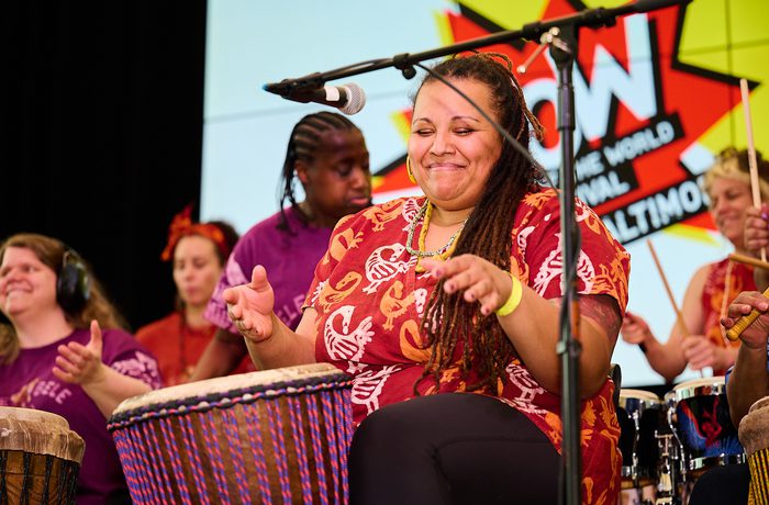 A group of diverse women joyfully play drums on stage. The central woman smiles with eyes closed. A banner in the background reads "WOW."