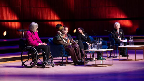 A panel of five women, one in a wheelchair, sit under warm lighting on a stage. The setting suggests a formal discussion or presentation.