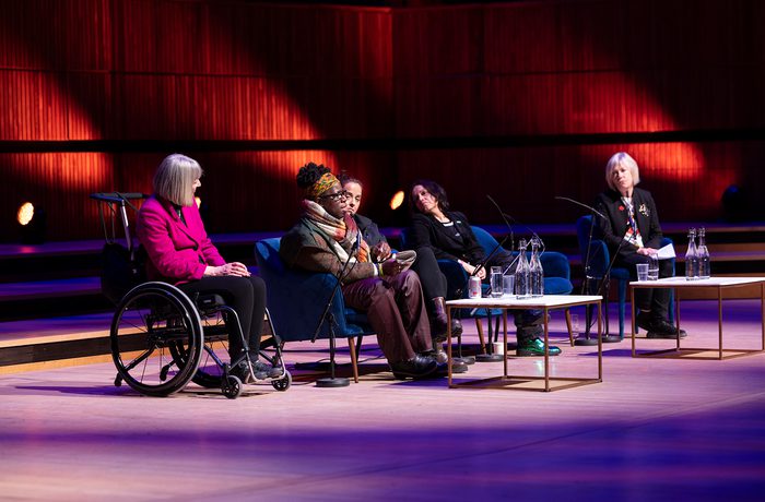 A panel of five women, one in a wheelchair, sit under warm lighting on a stage. The setting suggests a formal discussion or presentation.