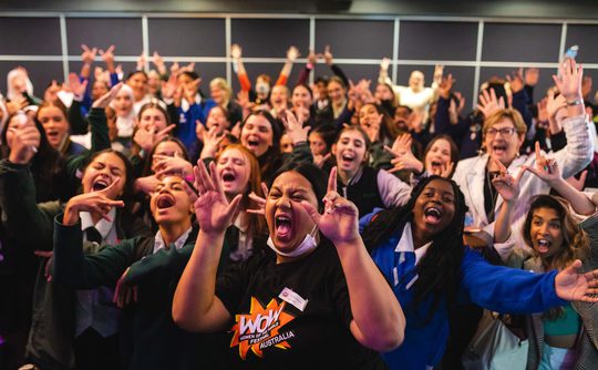 A diverse group of people excitedly pose together indoors, many smiling and raising their hands in joy. The mood is energetic and celebratory.