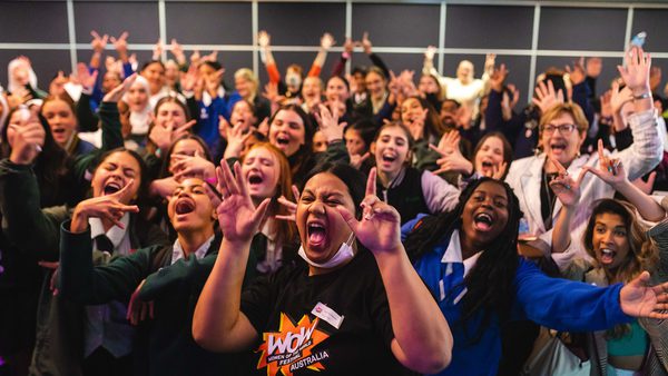 A diverse group of people excitedly pose together indoors, many smiling and raising their hands in joy. The mood is energetic and celebratory.