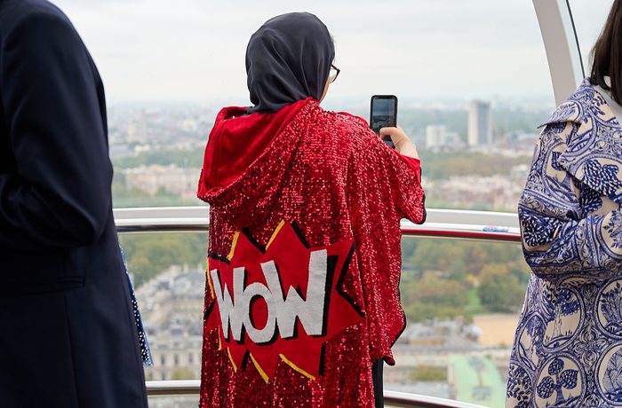 A person in a sequinned red cape with "WOW" on the back takes a photo from a Ferris wheel, overlooking a cityscape with an overcast sky.