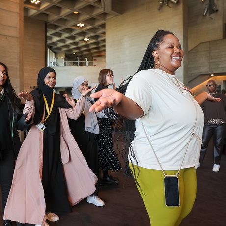 A group of diverse individuals joyfully dance in a modern building with a concrete interior.