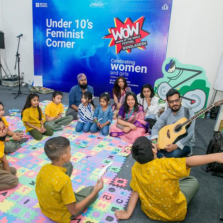 Children sit in a circle on colourful mats at the "Under 10’s Feminist Corner." A man plays guitar, with a vibrant wall display celebrating women.