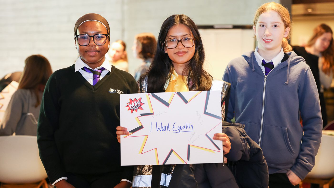 Three students stand together, smiling. The student in the center holds a sign that reads, "I Want Equality."