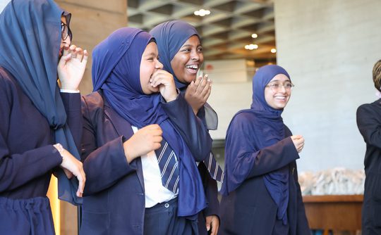 Four women wearing dark blue hijabs and uniforms laugh joyfully together indoors.