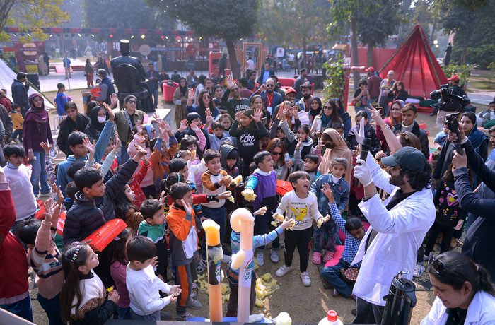 A scientist in a lab coat performs an experiment for excited children at an outdoor science fair, surrounded by onlookers and cameras, creating a lively atmosphere.